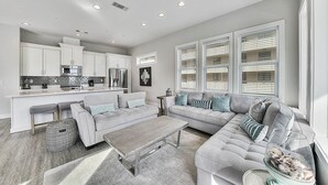 A modern living room with a grey sofa, a coffee table, and a kitchen area with white cabinets and appliances. - Aria Del Mare - Beach House with Ocean Views & Private Pool Area (Miramar Beach)