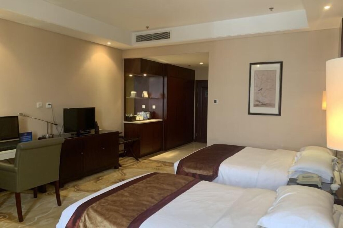 Quarto de hotel com duas camas, uma escrivaninha, televisão, banheiro e um quadro pendurado na parede.