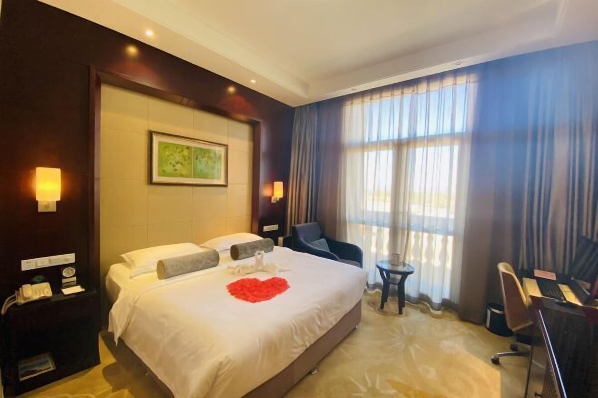Quarto de hotel com uma cama grande, uma escrivaninha com cadeira, uma mesinha pequena e uma mesa de cabeceira com telefone e abajur.