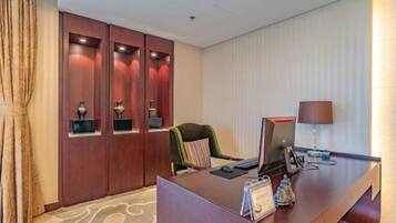 Zona de recepción de un hotel con escritorio, computadora y una silla verde. Se observan armarios de madera con objetos decorativos y una cortina a la izquierda.