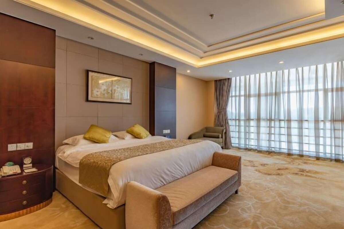 Quarto de hotel com uma cama grande, uma mesinha de cabeceira, uma cadeira e uma janela ampla com cortinas.