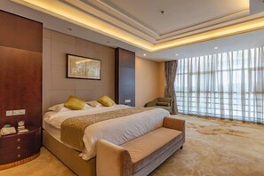 Quarto de hotel com uma cama grande, uma mesinha de cabeceira, uma cadeira e uma janela ampla com cortinas.