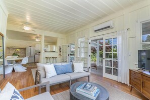 A spacious living area with a sofa, coffee table, and TV stand. There is a kitchen in the background and a sliding glass door leading to an outdoor area. - Kuuipo Hale on Weke Rd in Hanalei - short walk to the beach & A/C! TVNC #1159 (Hanalei)