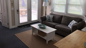 A living room with a brown sofa, a white coffee table, and a wooden table. There are two windows with white curtains and a door with a white frame. - Steps to the beach with an ocean view,  (Barnstable)