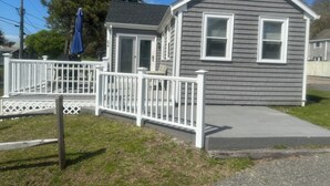A small, single-story house with a gray exterior and white trim. It features a front porch with a white railing and a blue umbrella. - Steps to the beach with an ocean view,  (Barnstable)