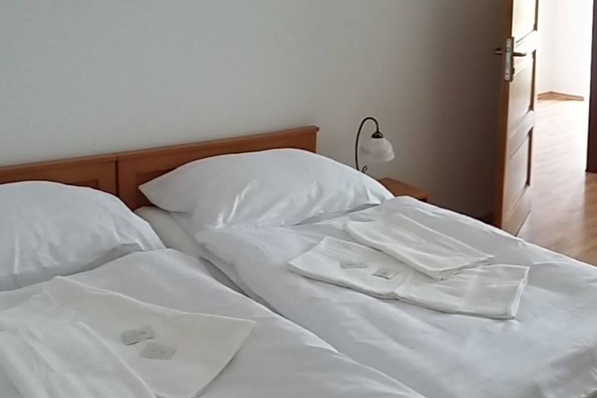 A double bed with white linens and a wooden headboard. There is a bedside table with a lamp on the left side.