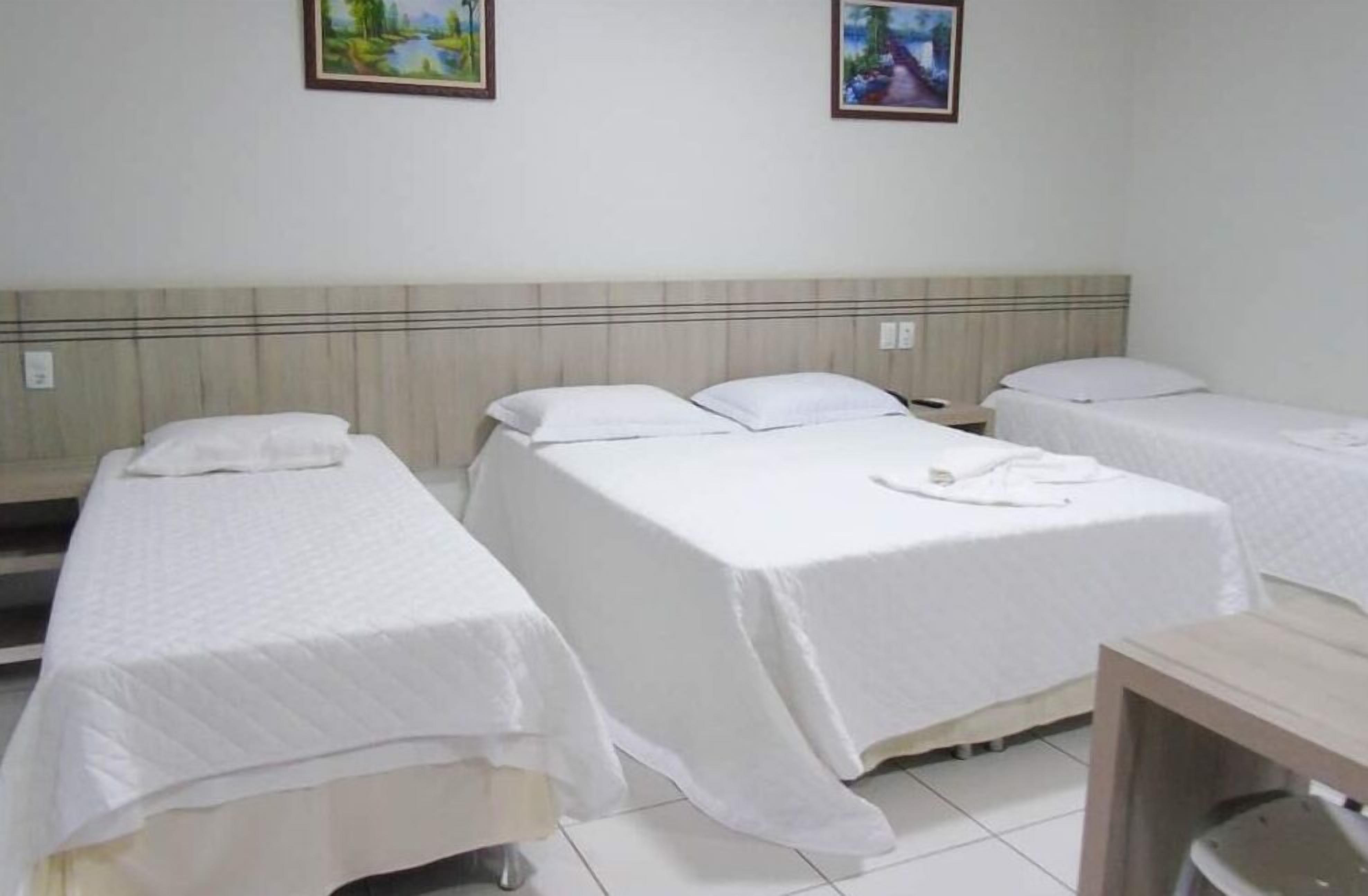 A hotel room with three single beds, each with a white bedspread and pillows. There are two framed pictures on the wall above the beds.