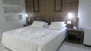 A neatly made bed with white linens and towels, a wooden headboard, a bedside table with a phone and a lamp, and a patterned wallpaper on the wall. - Maison Hotel (Tangará da Serra)