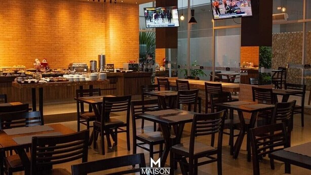 A modern dining area with wooden tables and chairs, a brick wall, and a television mounted on the wall. - Maison Hotel (Tangará da Serra)