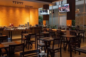 A modern dining area with wooden tables and chairs, a brick wall, and a television mounted on the wall. - Maison Hotel (Tangará da Serra)
