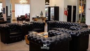 A hotel lobby with black leather sofas, a chandelier, a minibar, and a reception desk. - Maison Hotel (Tangará da Serra)