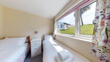 A bedroom with two single beds, a bedside table with a lamp, and a window with a view of a house and greenery.