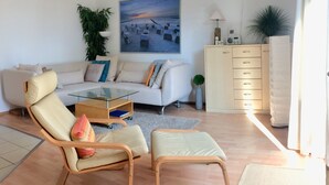Una sala de estar moderna con un sofá, una mesa de centro de vidrio, una silla, un mueble aparador y un cuadro de una playa.