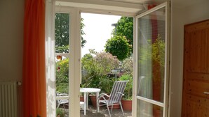 Puerta corredera de vidrio abierta que da a un patio exterior con una mesa y sillas, rodeado de plantas en macetas y una puerta de madera.
