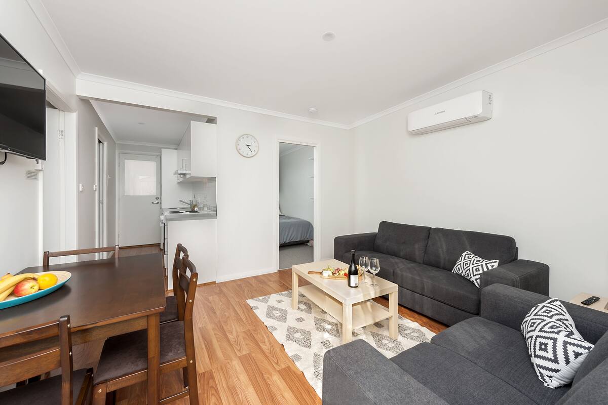 A modern living room with a grey sofa, wooden dining table, and a small kitchen area in the background.
