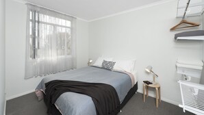 A bedroom with a bed, a bedside table with a lamp, a hanging clothes rack, and a window with sheer curtains. - 125 Milton (Milton)