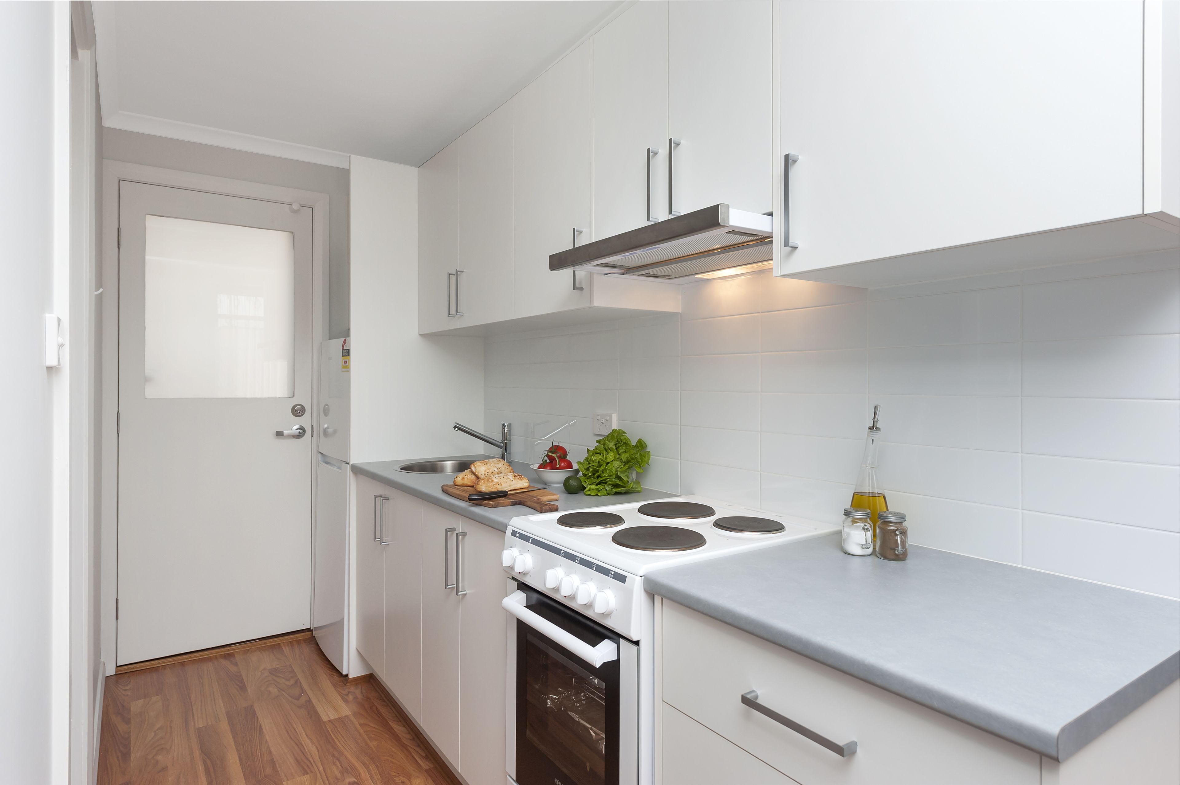 Una cucina compatta con mobili bianchi, forno integrato, piano cottura e un piccolo bancone con tagliere, verdure e condimenti.