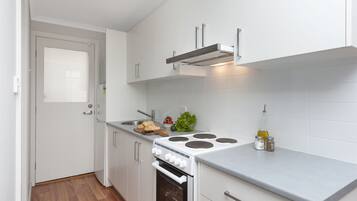 Una cucina compatta con mobili bianchi, forno integrato, piano cottura e un piccolo bancone con tagliere, verdure e condimenti.