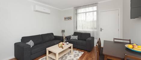 Un salon moderne avec deux canapés sombres, une table basse en bois, une table à manger avec des chaises et un téléviseur fixé au mur.