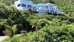 Un immeuble moderne à plusieurs étages, doté de grandes fenêtres et de balcons, entouré d’une végétation luxuriante et situé au pied d’une colline.