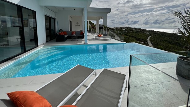 Un espace piscine extérieur moderne, doté d’une balustrade en verre, d’un coin salon avec des coussins et offrant une vue sur le paysage.