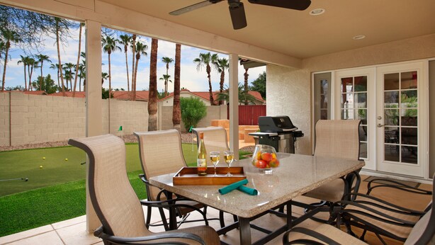 A patio with a table set for dining, chairs, a ceiling fan, and a view of a backyard with a lawn and palm trees. - Windrose McCormick Ranch (Scottsdale)