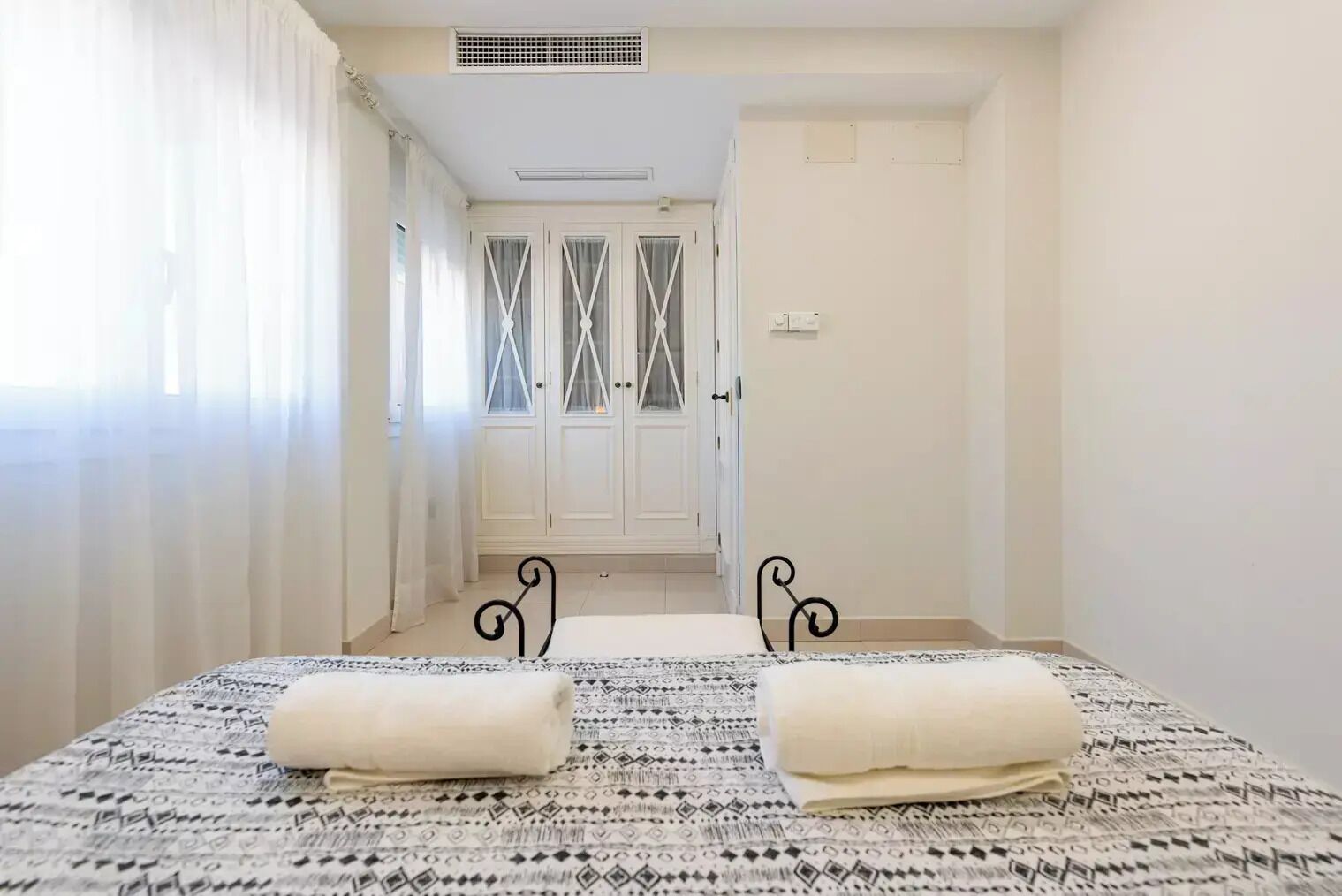 A bedroom with a bed, two pillows, a patterned bedspread, a door with a mirror, and a window with curtains.