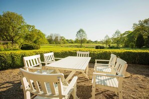 Een terras met een tafel en stoelen, omgeven door een verzorgde haag en uitzicht op een groen veld.