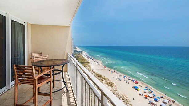 A balcony with a table and chairs overlooking a beach with umbrellas and people.