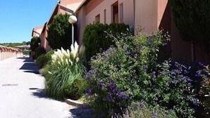 A building with a row of windows, a paved pathway, and various plants and shrubs. - T2 by the sea, 150 m to the beach/sea, TOPP prices, FREE: WLAN, pool, tennis (Le Pradet)