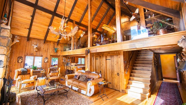 Un intérieur spacieux de chalet en bois, avec un plafond haut, une cheminée, un coin repas et un escalier menant à un étage supérieur.