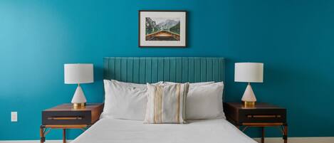 A neatly made bed with white linens, two bedside tables with lamps, a teal-colored headboard, and a framed picture of a mountain landscape on the wall.