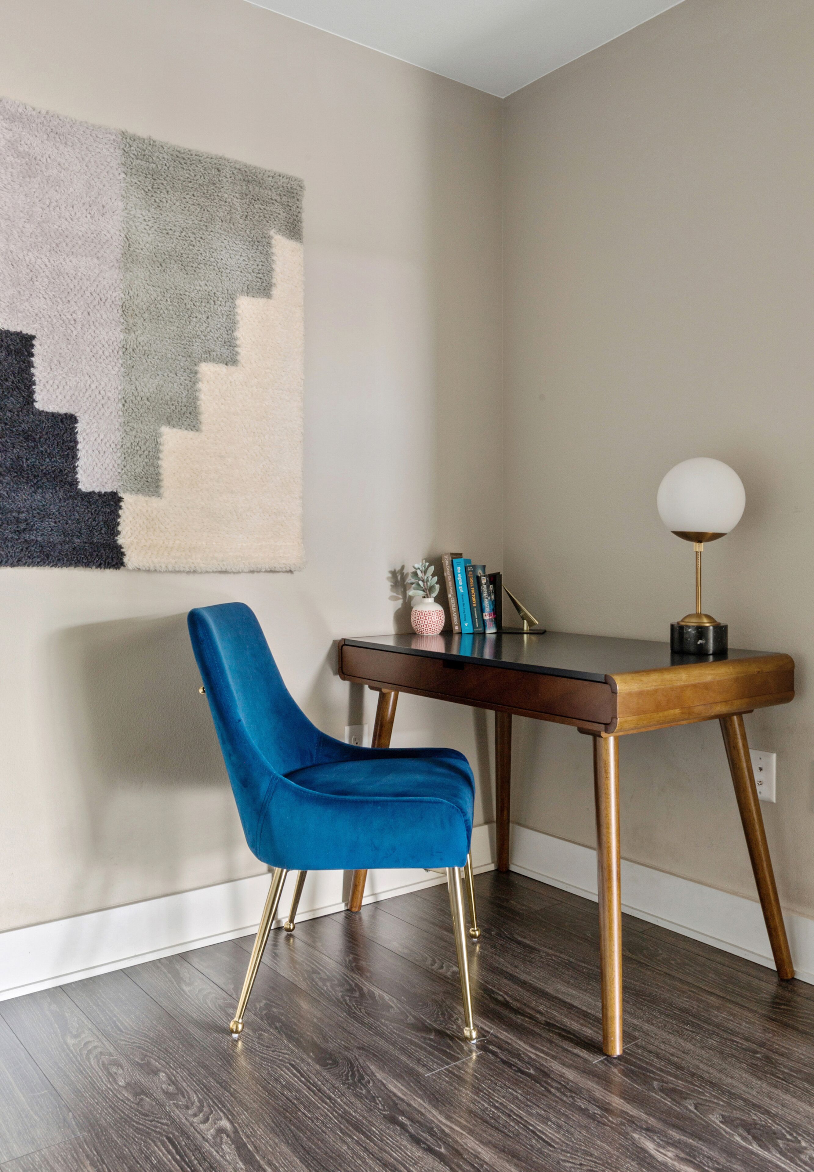 A room with a blue chair, a desk with a lamp, and a wall-mounted abstract art piece.
