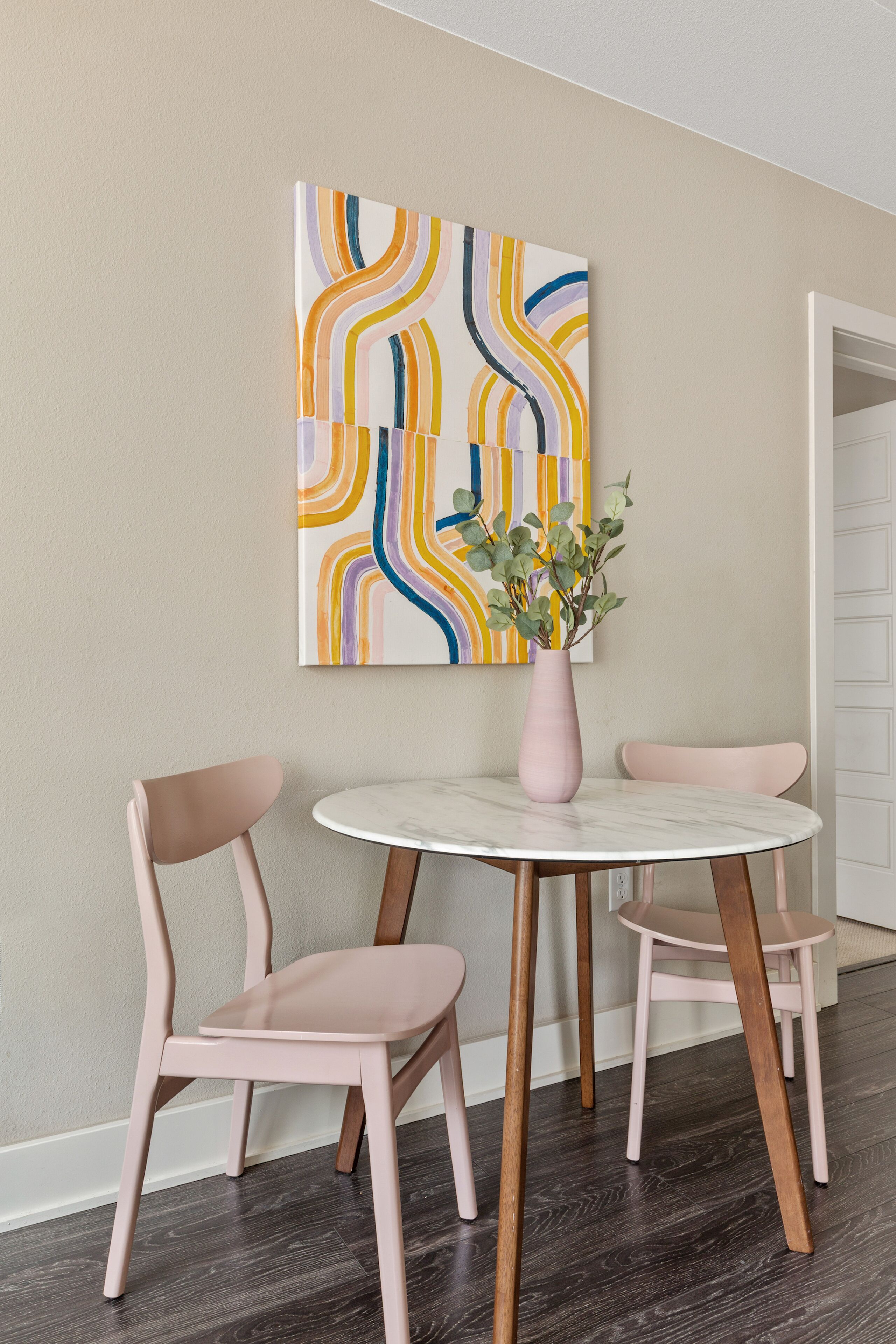 Un comedor con una mesa redonda, dos sillas, un florero con flores y un cuadro abstracto colorido en la pared.