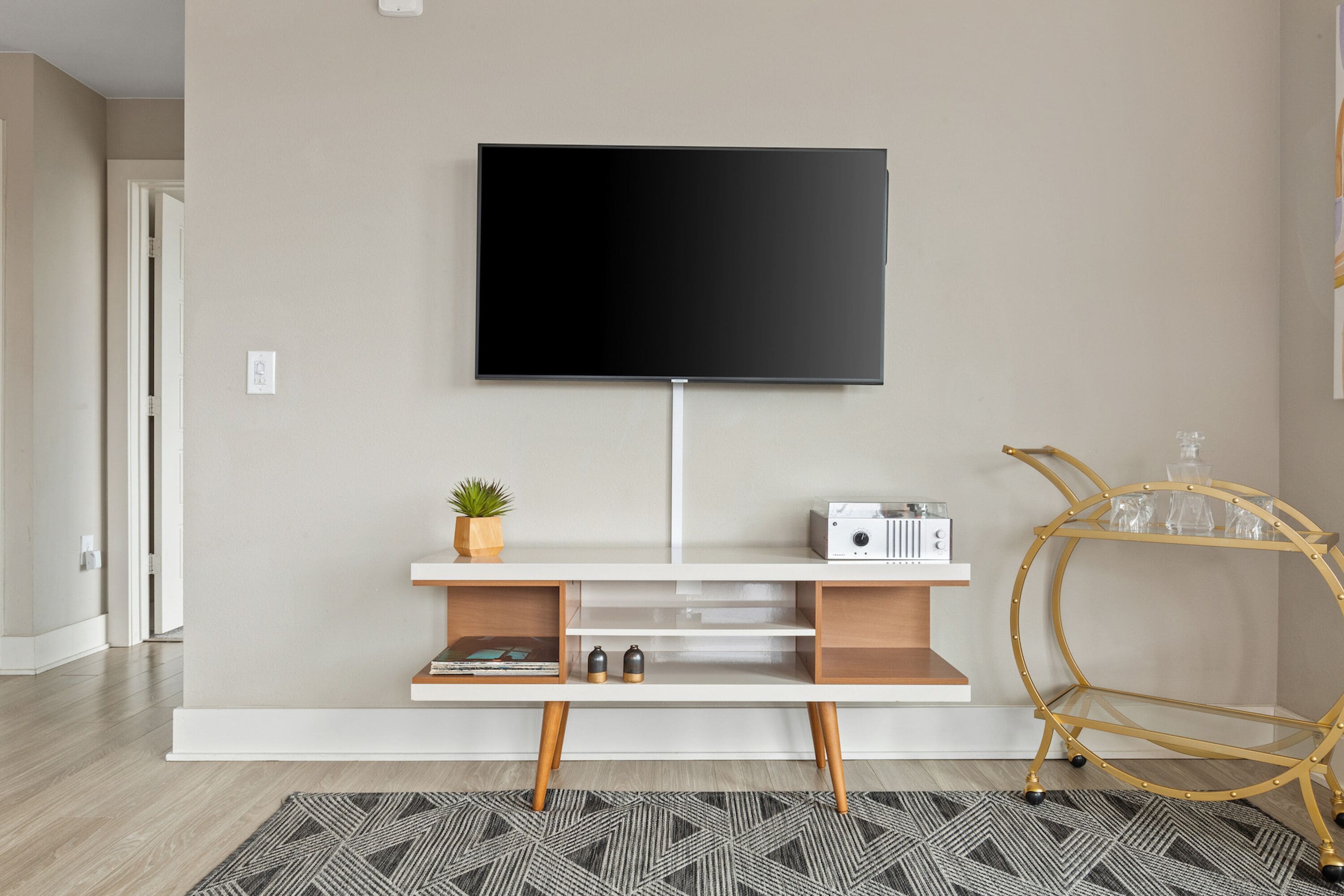 A modern living room with a flat-screen TV mounted on a white console table, a small plant, a radio, and a gold-framed side table with glassware.