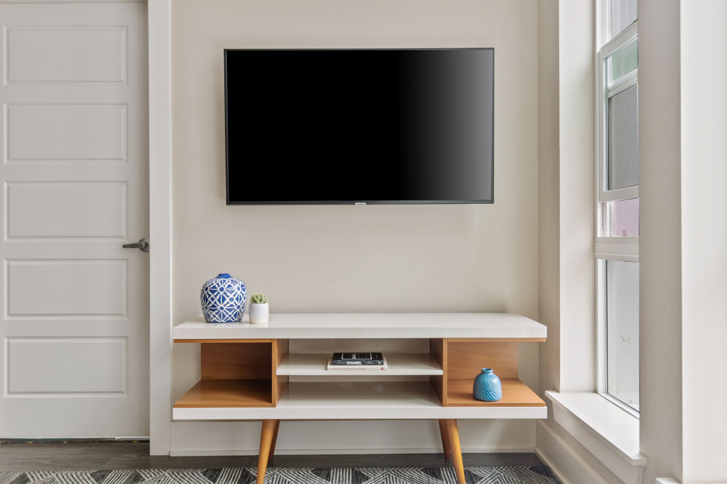 A modern living room with a flat-screen TV mounted on the wall, a white console table with shelves, and a window with a view.