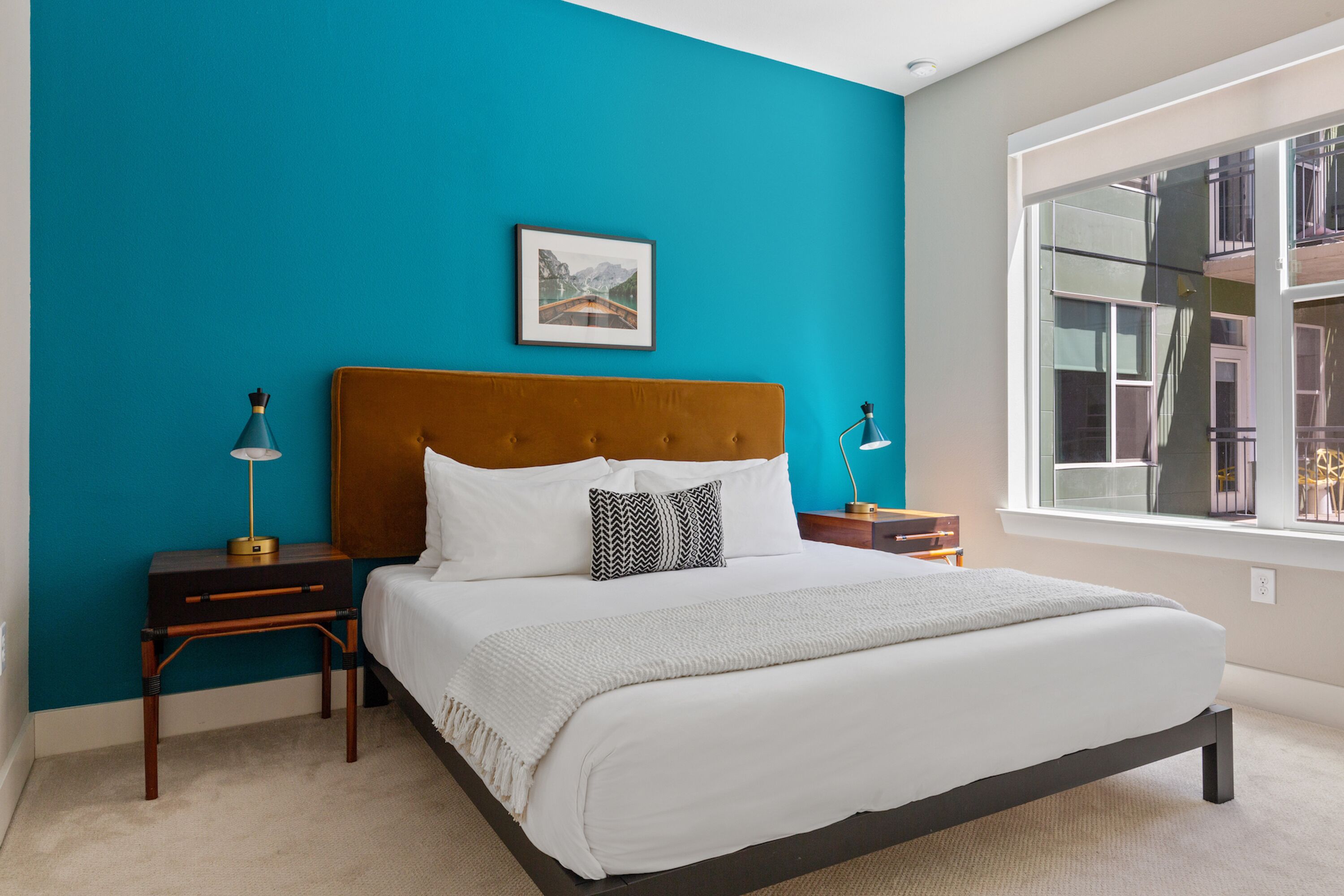A bedroom with a large bed, two bedside tables, a nightstand, a window with a view of buildings, and a picture on the wall.