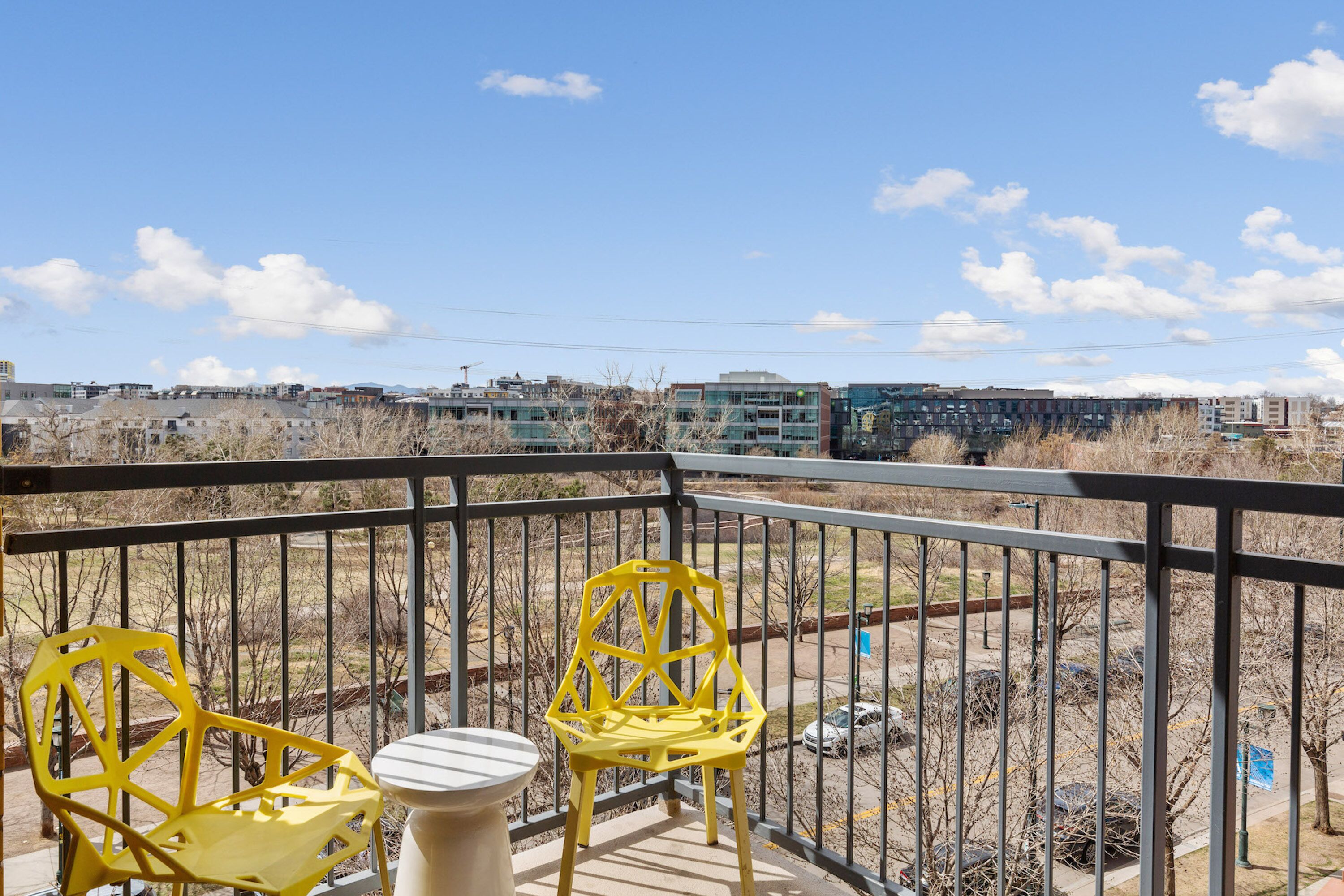 A balcony with two yellow chairs and a small table, overlooking a parking area and buildings in the distance.