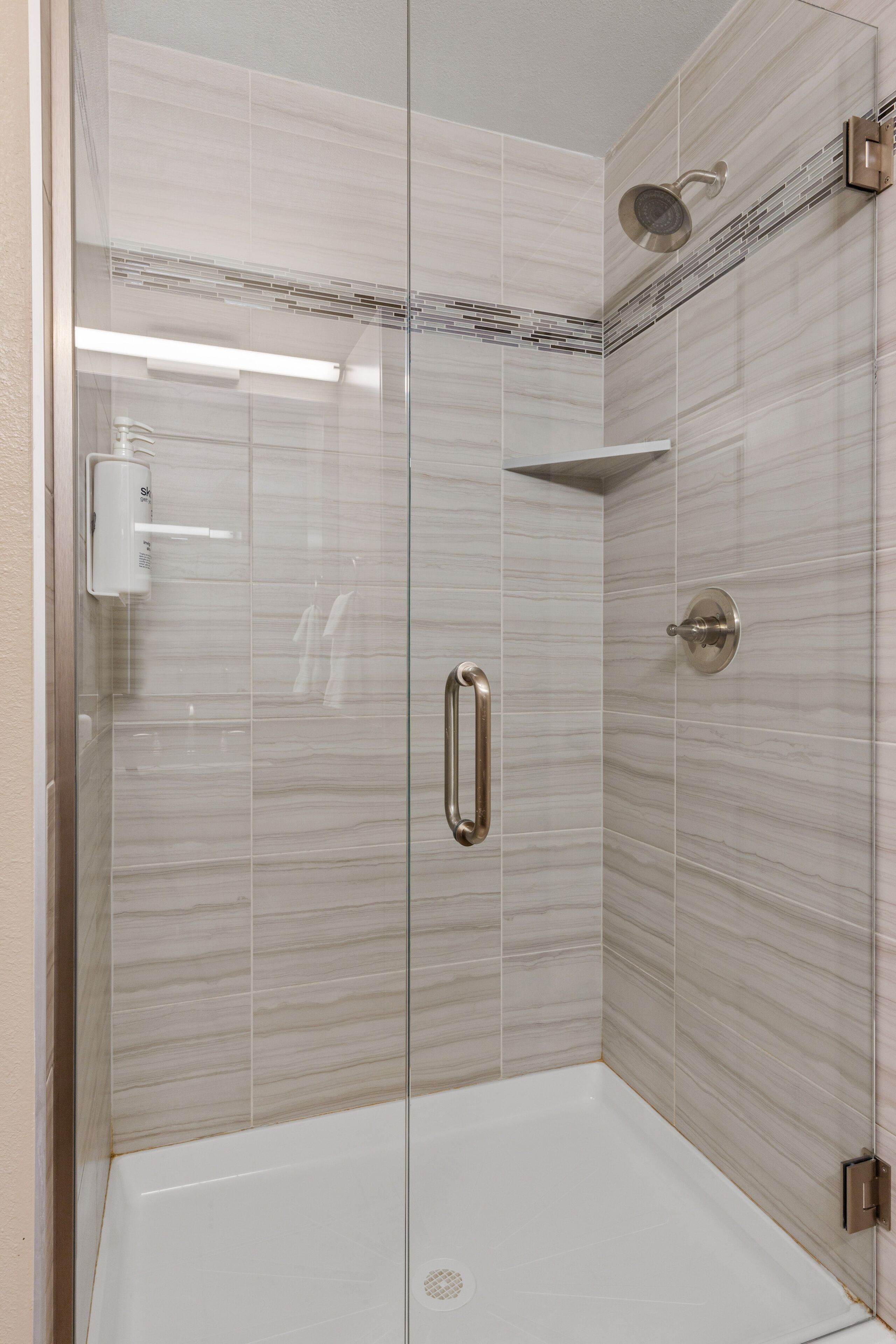 A glass-enclosed shower stall with a white tiled wall, a handheld showerhead, and a wall-mounted faucet.