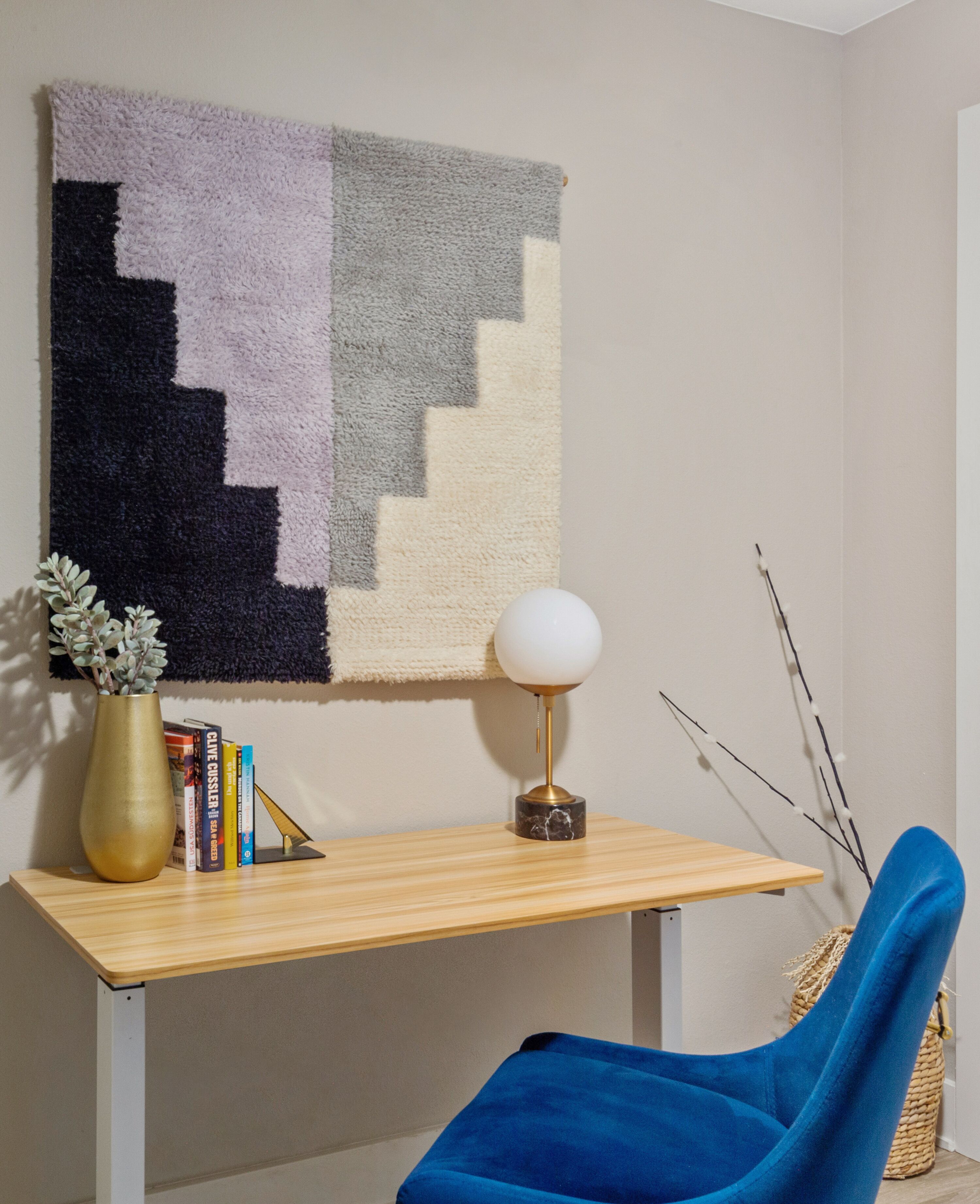 A room with a wooden desk, a blue chair, a wall-mounted lamp, a vase with plants, and a wall hanging.