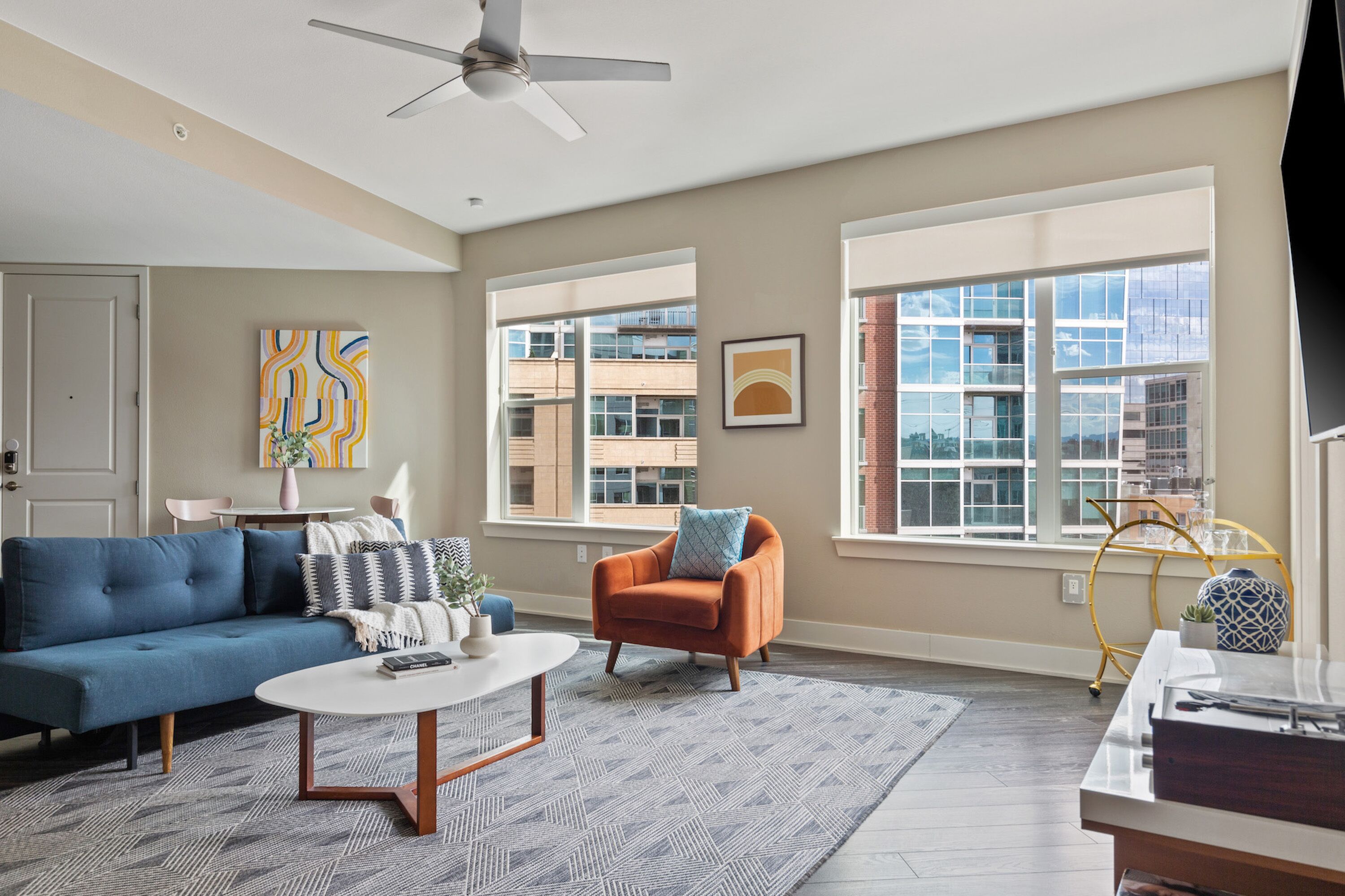 A modern living room with a blue sofa, an orange armchair, a white coffee table, and a large window with a city view.