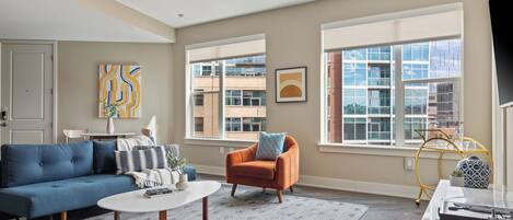 A modern living room with a blue sofa, an orange armchair, a white coffee table, and a large window with a city view.