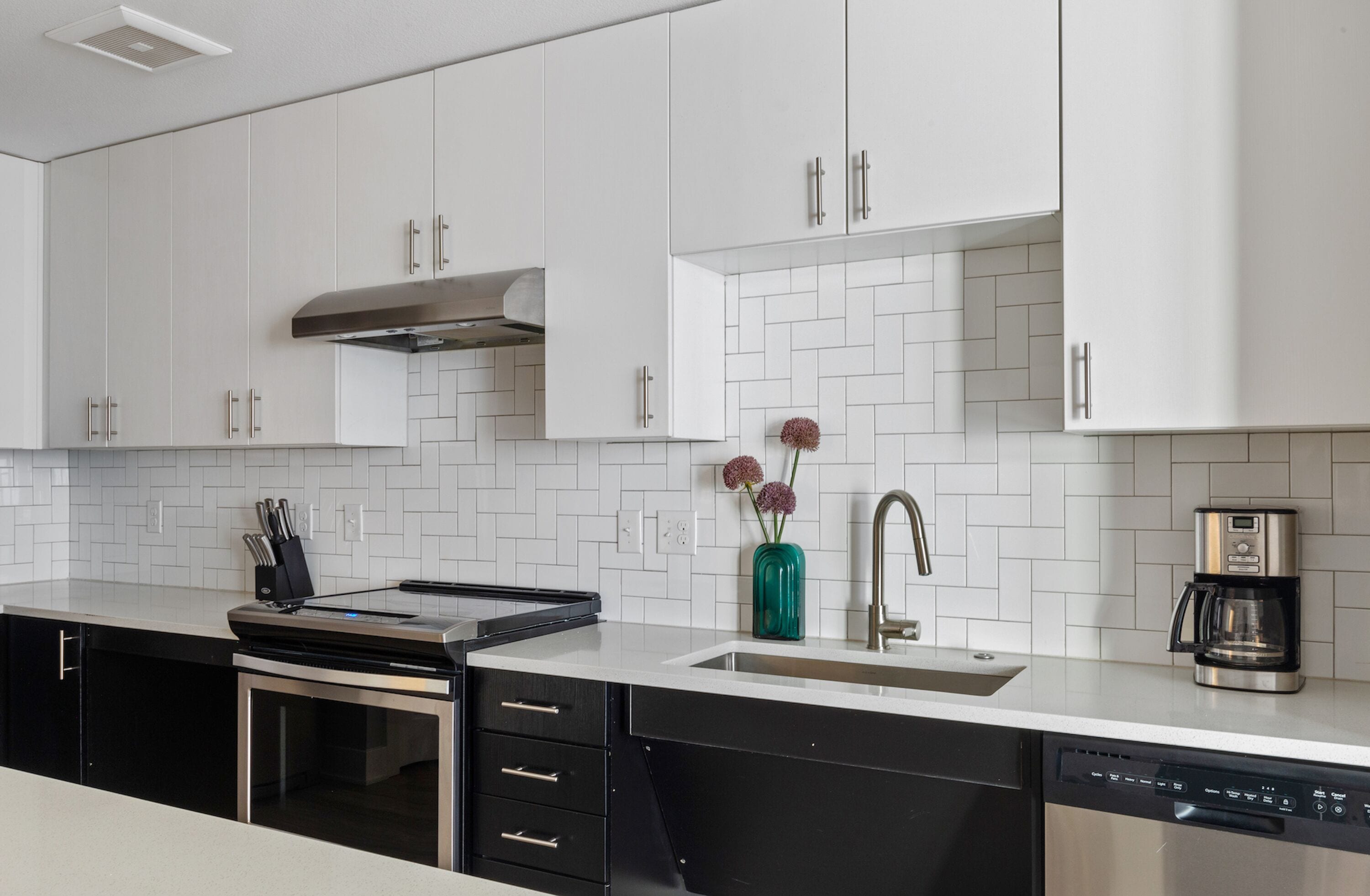 A modern kitchen with white cabinets, a stainless steel range hood, a dishwasher, and a coffee maker.