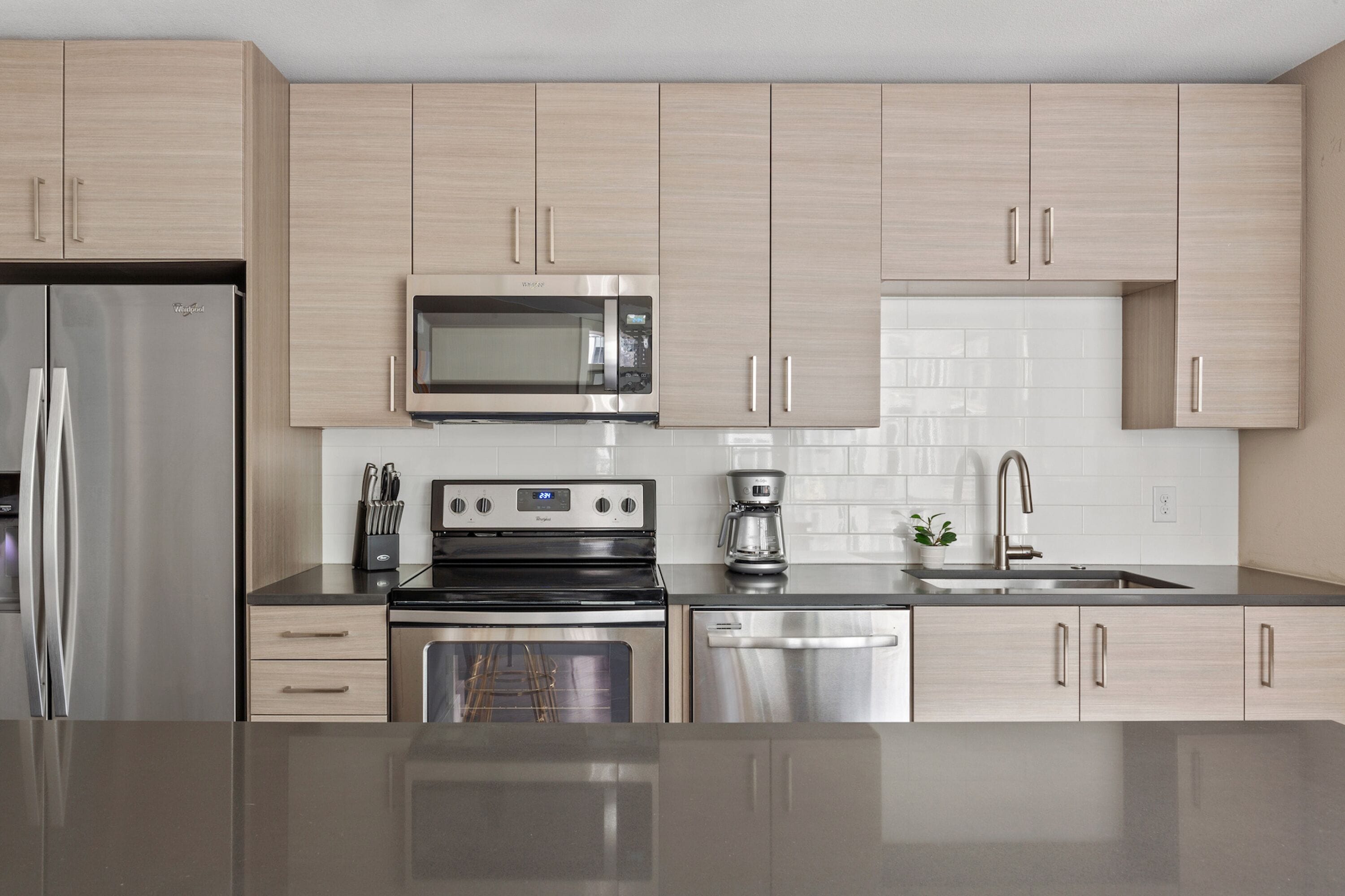 A modern kitchen with stainless steel appliances, light wood cabinets, and a white tiled backsplash.