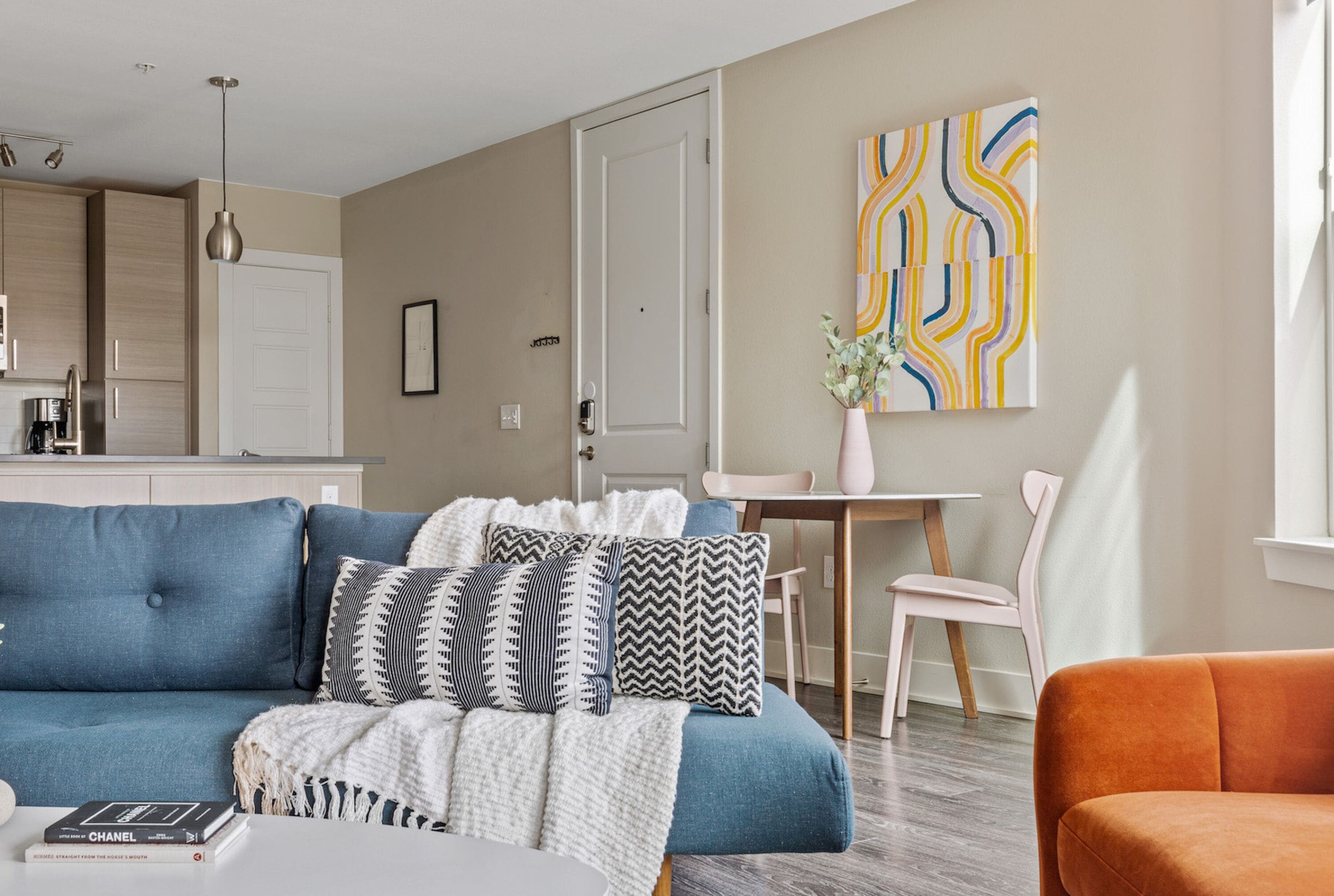 A modern living room with a blue sofa, a white coffee table, and a pink chair. There is a dining table with chairs and a vase of flowers. The room has a neutral color scheme with beige walls and a wooden floor.