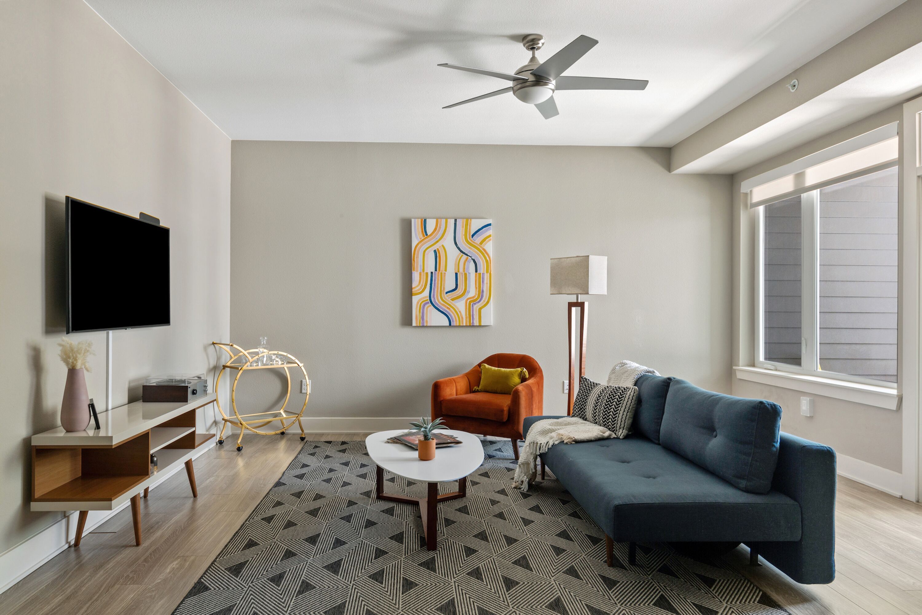 Un salon moderne avec un téléviseur à écran plat, un canapé bleu, un fauteuil orange, une table basse blanche et un tapis à motifs.