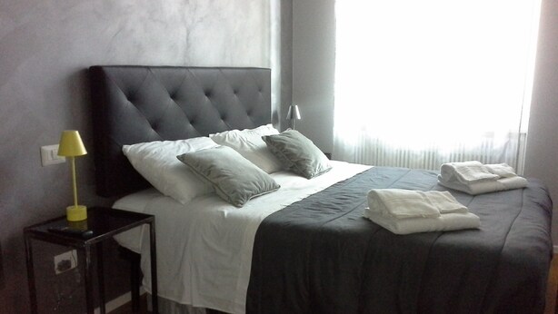 Una cama bien hecha con un cabecero oscuro, ropa de cama blanca y dos almohadas. Hay una mesita de noche con una lámpara y una toalla doblada sobre la cama.