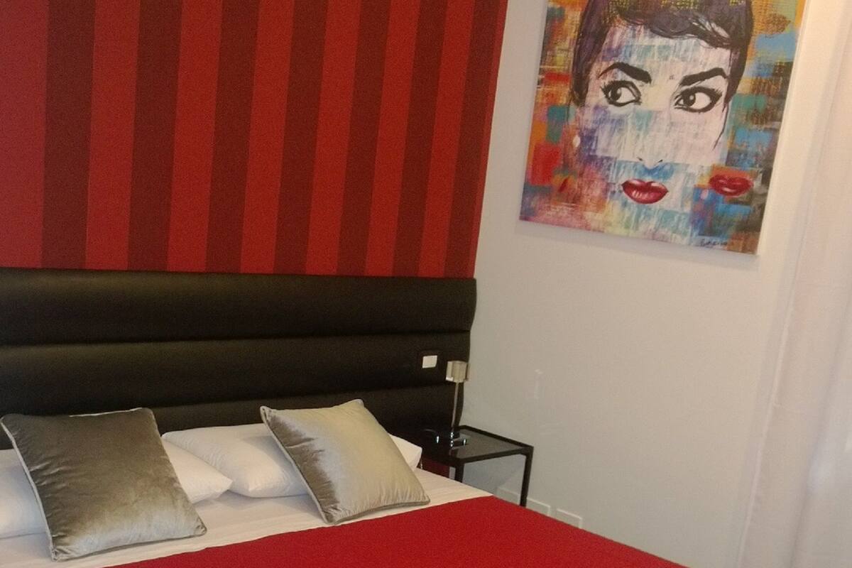 Une chambre à coucher avec une couvre-lit rouge, une tête de lit noire et un tableau accroché au mur.