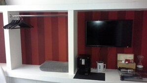 Una habitación compacta con una cama, un televisor de pantalla plana en la pared, una cafetera y una mesita con una lámpara y libros.