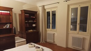 A hotel room with a bed, a wooden bookshelf, a radiator, and a window with curtains.
